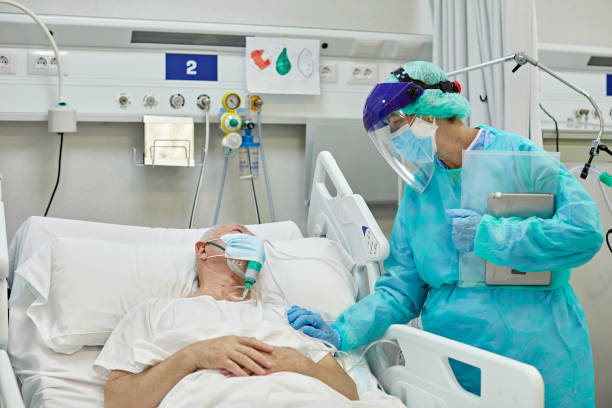 Female Doctor Supporting Male Patient Lying in Hospital Bed Mature female medical professional wearing protective garment, face shield, surgical mask, and gloves consoling senior male patient recovering from coronavirus. frontline worker mask stock pictures, royalty-free photos & images