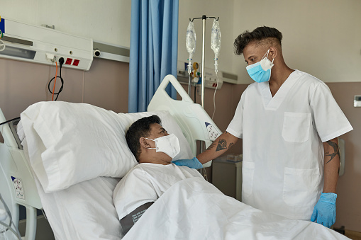 Young male nurse in white scrubs standing at bedside with hand on shoulder of recovering mature male COVID-19 patient, both wearing protective masks.