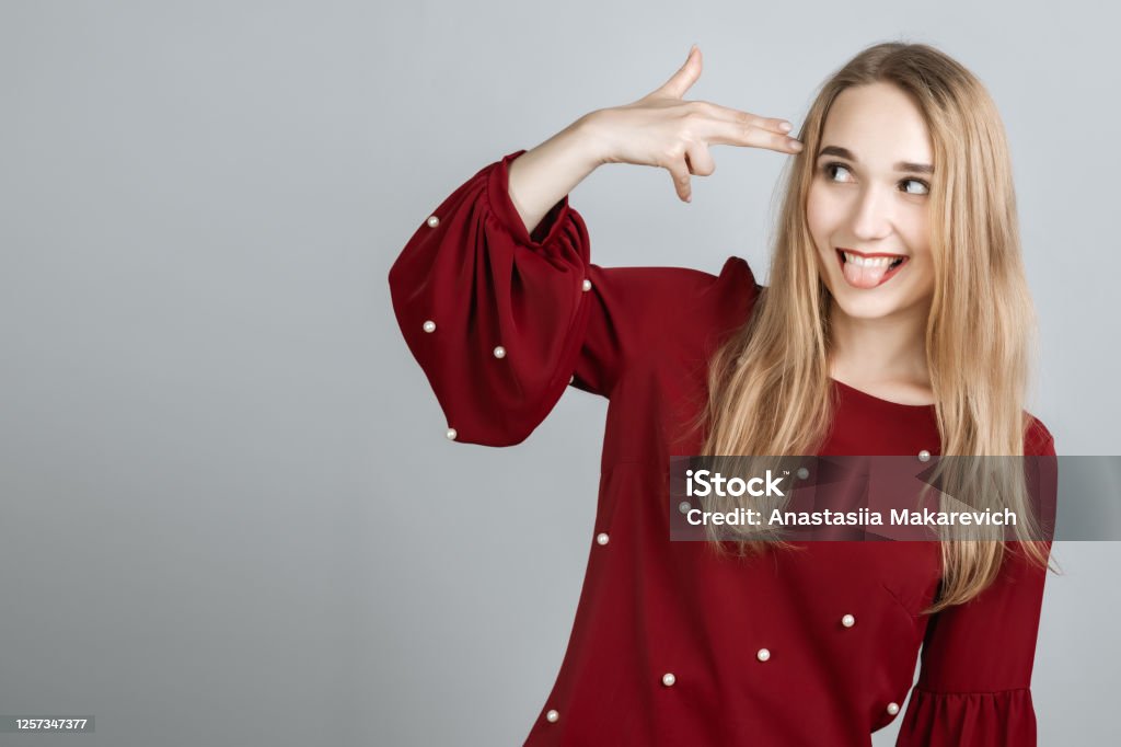 I'm fed up with this. Stylish European girl with long blonde hair shoots in temple, tilts head, dressed in casual t shirt, demonstrates suicide gesture, isolated on gray background Adult Stock Photo
