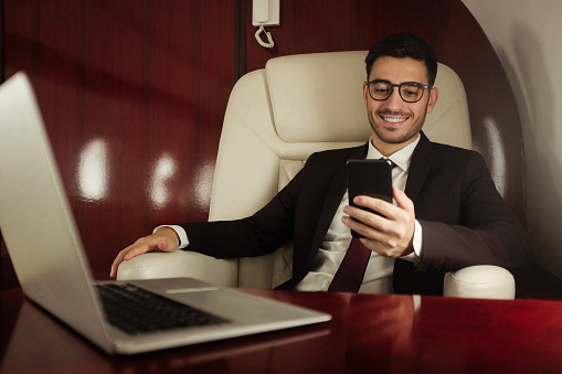 Handsome businessman travelling first class, sitting in armchair next to window, reading text messages on phone screen