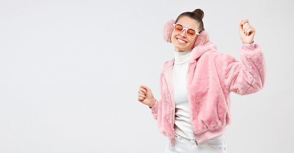 Horizontal banner of young female dancing in pink fluffy bomber, colored glasses and ear muffs, feeling happy at party, isolated on gray background