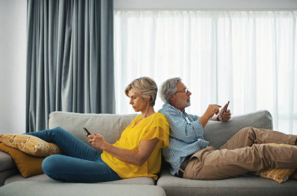 Middle aged couple having a morning coffee on sofa and browsing their phones. Closeup side view of a cheerful mid 50's couple having a morning coffee on a living room sofa and browsing their social media on their phones.   They are enjoying their weekend at home during coronavirus quarantine. mid adult men stock pictures, royalty-free photos & images