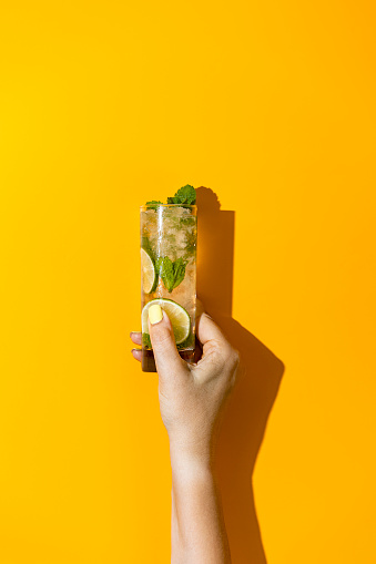 Vaso de mano de mujer con gotas de agua de bebida de cóctel Mojito con hielo de lima y menta sobre fondo amarillo brillante. Concepto de comida artística de verano sobre fondo amarillo. photo