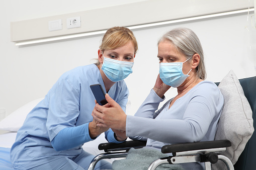 nurse helps with cell phone to contact the elderly lady's family in the wheelchair, wearing surgical protective medical masks in hospital room, concept of isolation from corona virus covid 19