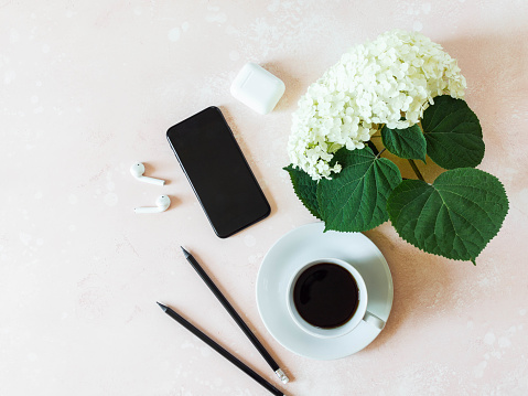 Home office desk workspace with black smartphone, black coffee in cup, white hydrangea, white earphones and graphite pencils on pink background. Flat lay, top view girl work business concept for lifestyle blog