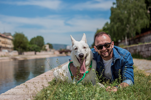 Young man and white dog lying in the grass by the river and posing for a picture. They are both looking at the camera and smiling