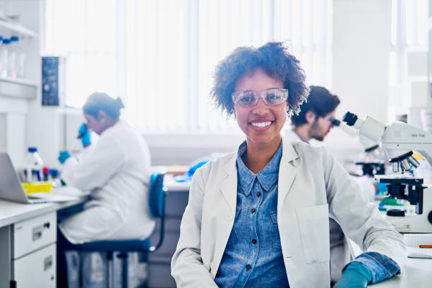Smiling young scientist working with her team in a lab Portrait of a smiling young female scientist sitting a table in a lab with colleagues working in the background african american scientist stock pictures, royalty-free photos & images