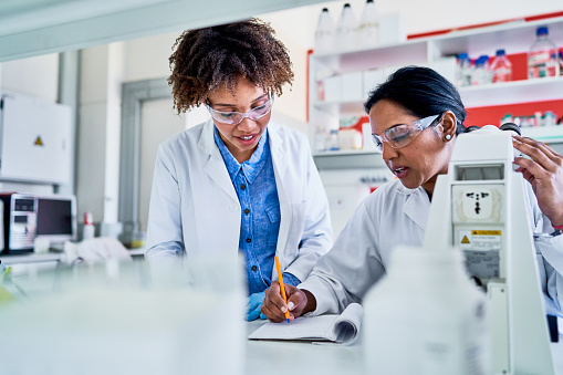 Two female scientists talking together and writing data in a notebook while working at a table in a lab
