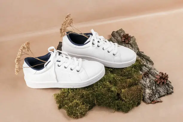 Photo of Ethical vegan shoes concept. A pair of white sneakers with dry flowers on tree bark and moss, neutral beige craft paper background
