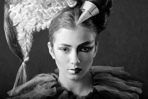 Beautiful Caucasian brunette close-up, with half face painted with white make-up  and half painted wih graphic black make-up. Her hair is half white with a wing made of braided hair and half black with a silver horn. She is loooking straight, in black and white. Angel and Devil Concept