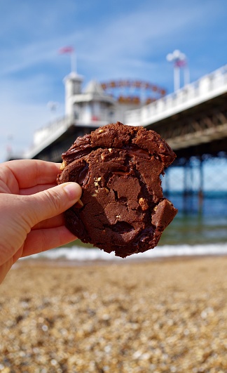 Vegan double chocolate chip cookie enjoyed on Brighton Beach, bought locally. The cookie is held up by a woman's hand with Brighton Palace Pier in the background. A vegan diet excludes all animal products. Many people choose to eat this way for ethical, environmental or health reasons.