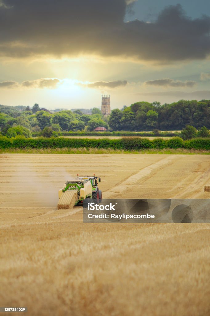 Straw or hay Baler machine pulled by a tractor, discharging a compacted straw bale from the rear in a field of recently harvested barley on the outskirts of Cirencester in the Cotswolds Agricultural Field Stock Photo