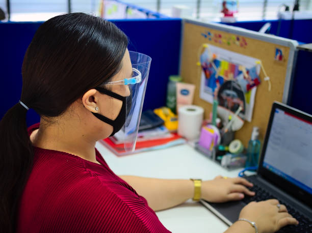 Woman wearing face mask while working in office A young, beautiful woman wearing a face mask as protection against covid-19 while working inside an office. office cubicle mask stock pictures, royalty-free photos & images