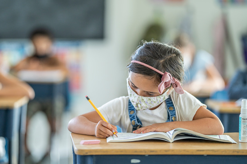 6 year old girl wearing a reusable protective face mask while working at her desk in school.
