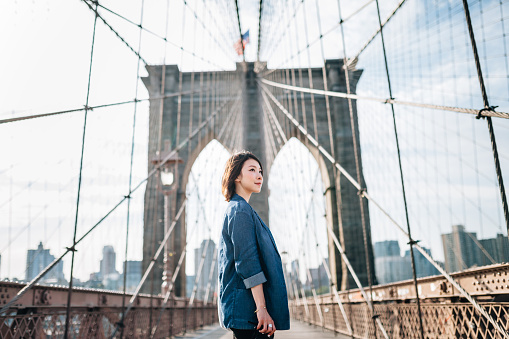 Low angel shot of an aspirational young Asian businesswoman on Brooklyn Bridge against New York City cityscape