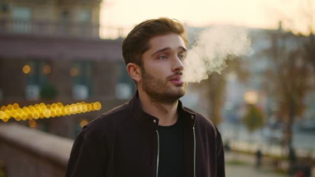 Serious guy smoking e-cigarette outdoors. Thoughtful man vaping on street.