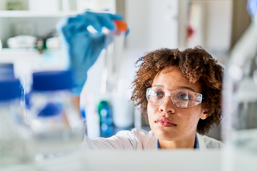 Young female scientist holding up a sample in a bottle while working in a lab full of research equipment