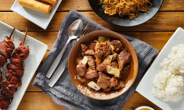 pork adobo meal with filipino foods such as lumpia, pancit noodles, and rice top down