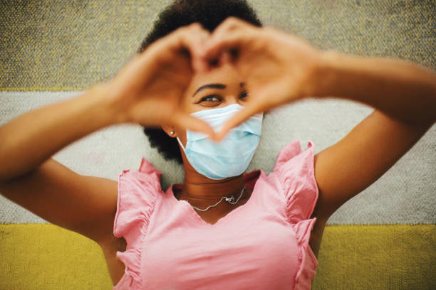 Smiling behind protective face mask. African american young woman lying on the floor, smiling behind protective face mask. Looking at camera through heart shape that she made with her hands. woman lying on the floor isolated stock pictures, royalty-free photos & images