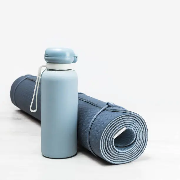 Rolled blue yoga mat and blue water bottle on grey wooden surface. Gender neutral fitness yoga and exercise concept with copy space. Active lifestyle. Workout at home or gym banner