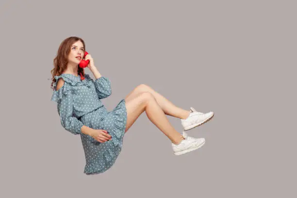 Charming girl in ruffle dress holding phone handset levitating, hovering floating air and having conversation on telephone, looking up with dreamy expression. studio shot isolated gray background,