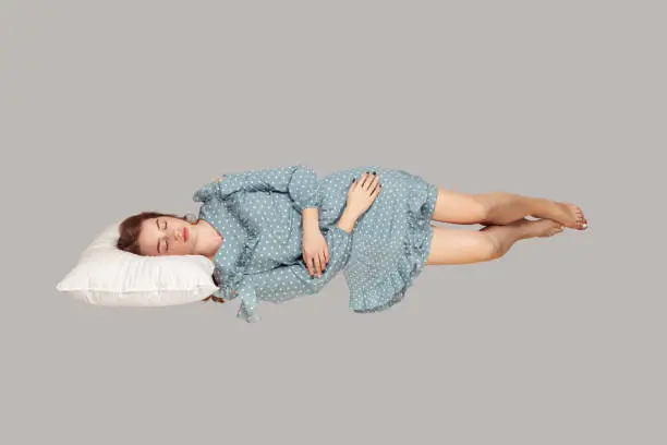 Photo of Sleeping beauty hovering in air. Relaxed girl in vintage ruffle dress lying comfortably on pillow levitating