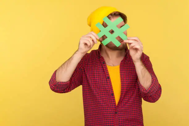 Photo of Hashtag, internet trends. Hipster guy in beanie hat, checkered shirt looking through big hash sign, curious watching