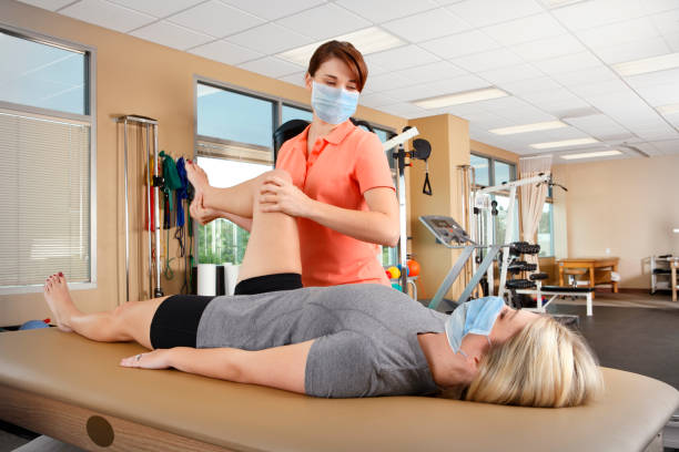 Physical Therapist Wearing Protective Face Mask Evaluates Range Of Motion A physical therapist, wearing a protective face mask, evaluates the range of motion of a young female patient's hip as she lies on a treatment table. The scene is inside a physical therapy clinic with several pieces of exercise equipment in the background.  The therapist is in her early thirties and the patient is in er early twenties. physical therapy recovery touching human knee stock pictures, royalty-free photos & images
