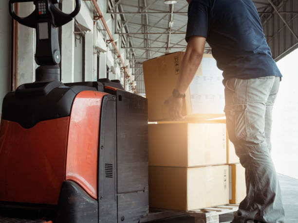 istock Warehouse worker lifting boxes on pallet with electric forklift pallet jack. Manufacturing warehouse shipment package boxes. 1257418921