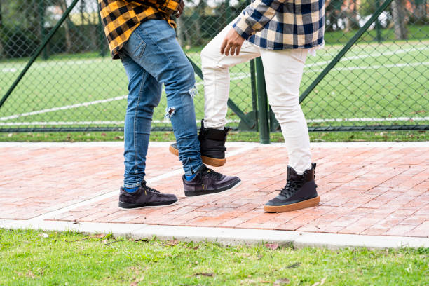 Latino friends men of approximately 30 years of age dressed in a plaid shirt and wearing their face masks on the street greet each other with foot to avoid contagion