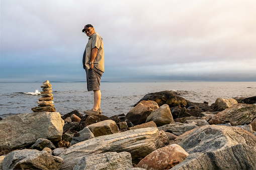 A man standing next to stone cairn on the ocean shore in Connecticut, USA, near Harkness Memorial State Park