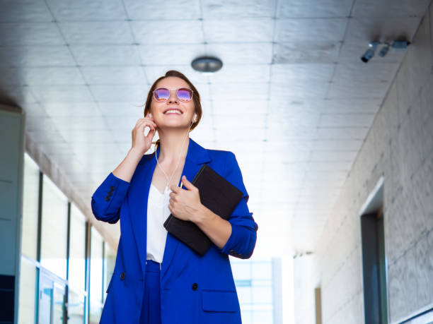 A woman in a business suit carries a folder with documents and smiles while talking on the phone via hands-free headset
