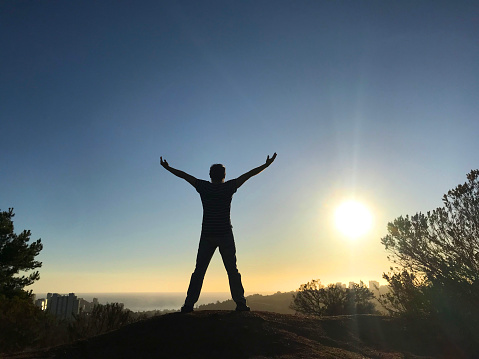 Young backpacker traveler hiker man celebrating and standing with arms outstretched on top of the mountains with a panoramic aerial landscape view of Pacific Ocean and sunset on back in Vina Viña del Mar, Reñaca, Valparaiso, Chile.