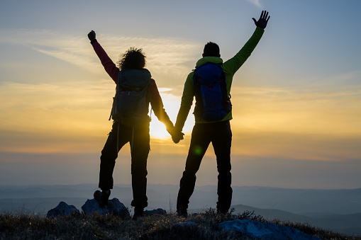 Rear view of a couple's silhouettes on top of a hill as they looks at the sun and valley with arms raised.