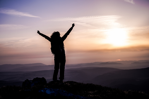 Rear view of a woman's silhouette on top of a hill as she looks at the sun and valley with arms raised.