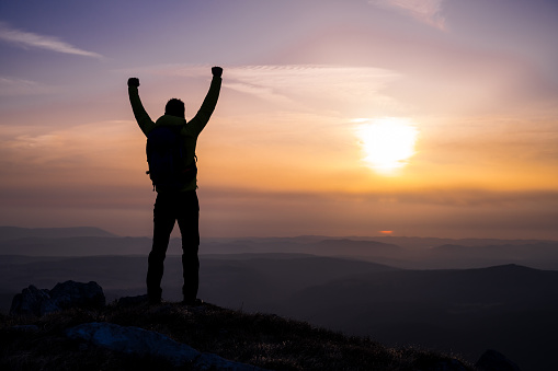 Rear view of a man's silhouette on top of a hill as he looks at the sun and valley with arms raised.