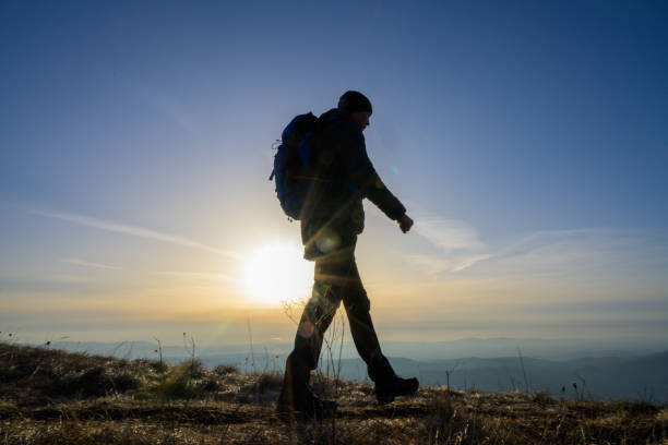 Silhouette of a man walking on a meadow on a hill. Sun and sky in the background.