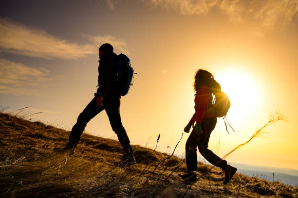 Photo of silhouettes of a hiker couple walking on top of a hill. Sun in the background.