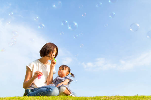 小さな女の子と母は青空に対してシャボン玉で遊ぶ - bubble wand child blowing asian ethnicity ストックフォトと画像