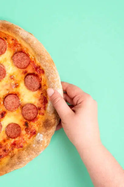 Vertical image with a woman's hand grabbing a pepperoni pizza. Top view image with a freshly baked pepperoni pizza on a green-mint background.