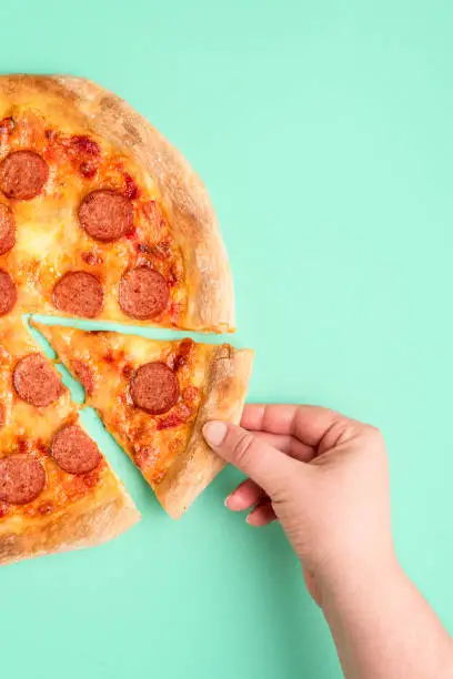 Woman's hand grabbing a slice of pepperoni pizza, top view. Pizza isolated on a green-mint background. Freshly baked pizza with salami, mozzarella and tomato sauce