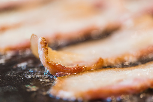 A close up photo of strips of bacon cooking on a grill.  One strip curls on the end as it cooks.