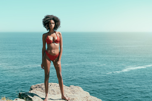 Dazzling young African-American female with a curly Afro hair and in a swimsuit is standing on a rock on a beach resort, horizon behind; with a copy space place on the right for a text advert message