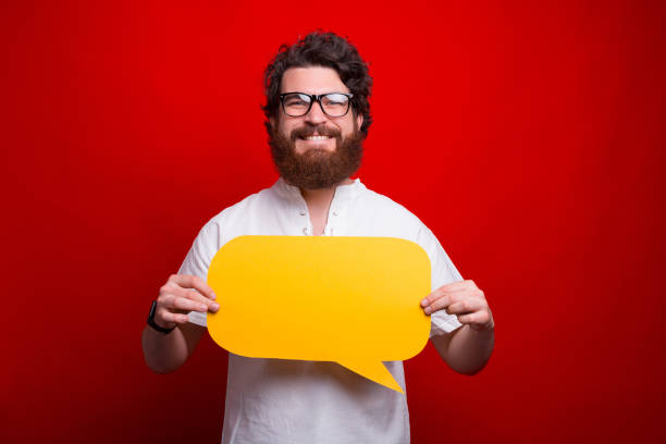istock Smiling bearded man wearing glasses is holding a yellow bubble speech on red background. 1257458095