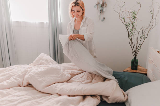 uma jovem bonita está fazendo uma cama. roupa de cama fresca. coloque a capa do travesseiro. sala leve e brilhante. bege, branco, cinza. fazendo melhorias em casa. limpeza e organização. casa aconchegante. morar em apartamentos. - sheet human hand bed women - fotografias e filmes do acervo