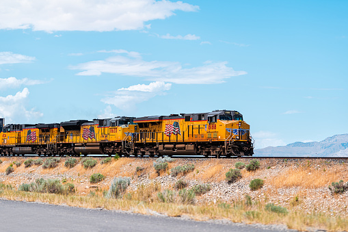 Salt Lake City, USA - July 27, 2019: Utah landscape with train on railroad with cars containers, and american flag on yellow color locomotive with mountains in desert background