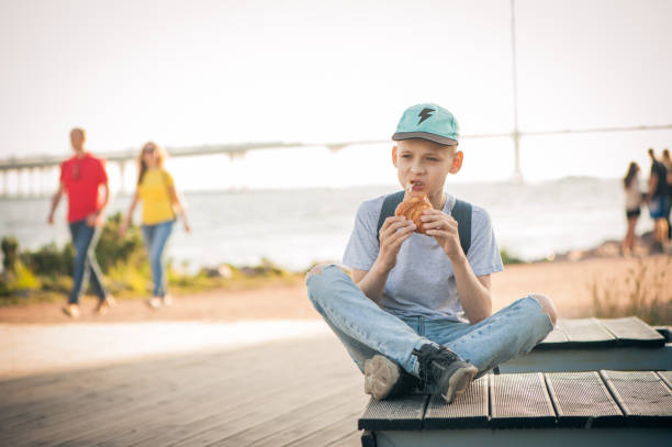 istock The boy sits on a bench on the waterfront with his legs crossed and eats a croissant. A teenager in the street eats pastries and looks thoughtfully into the distance. People are passing by. 1257462015