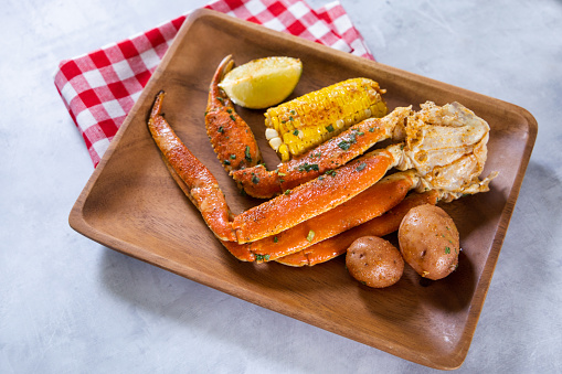 a wood tray full of seafood including garlic Alaskan crab legs, corn on the cob, new potatoes and a lime with a red picnic napkin
