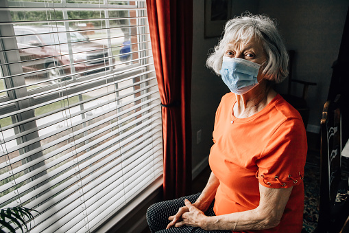 isolated elderly woman sitting near the window at her home or a nursing home with a mask on during quarantine