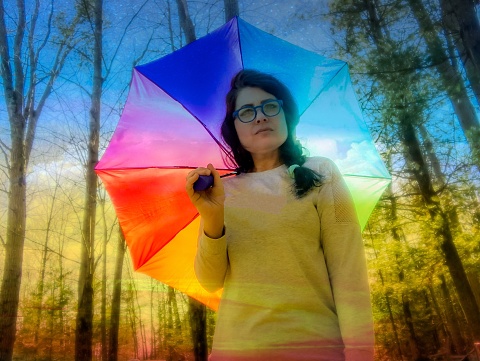 Beautiful woman standing outdoors in nature holding a colorful umbrella and looking proudly at the camera. The photograph has a dream-like feel to it with digital enhancements added in post production.